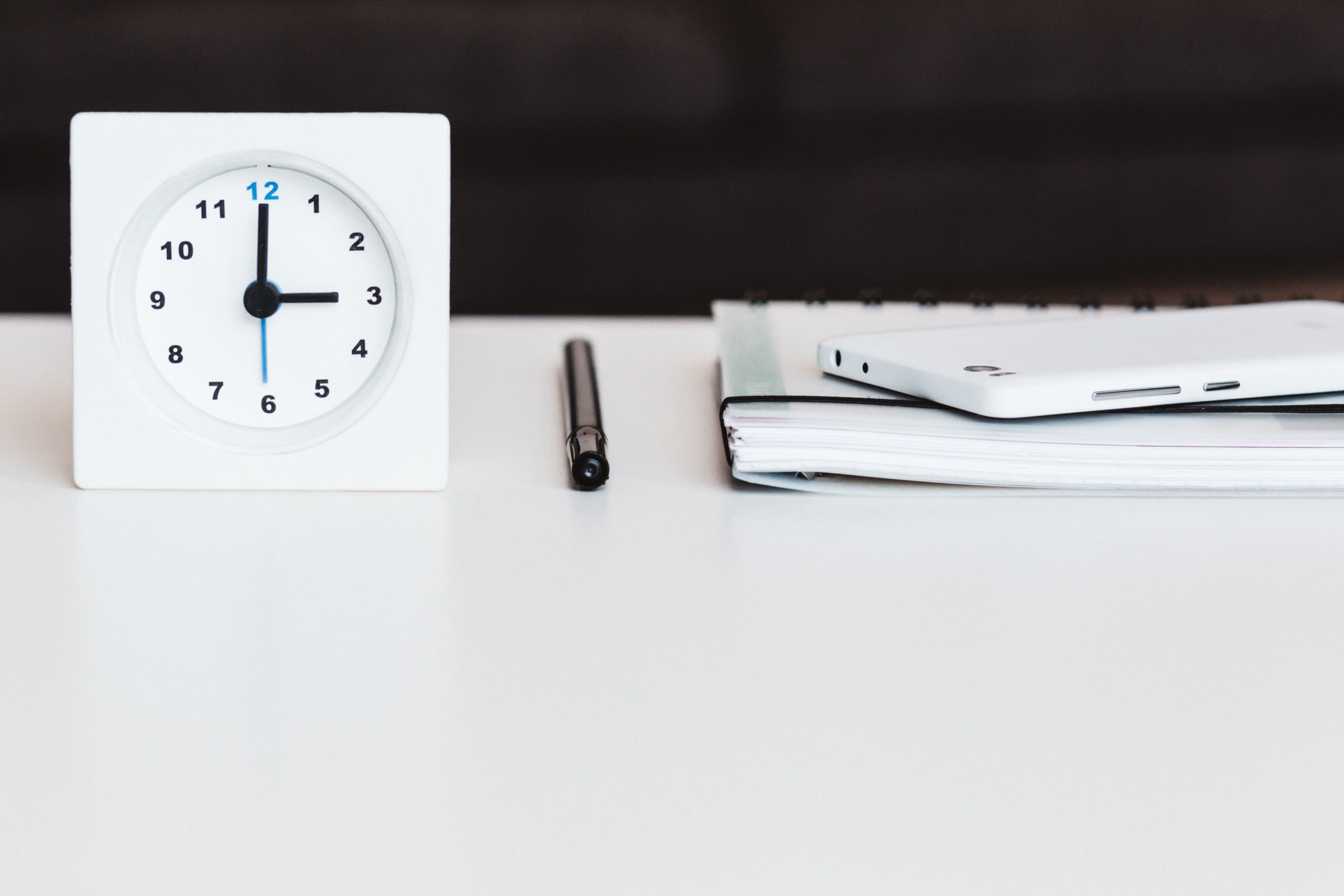 A clock, pen, notebook, and smart phone on a white desk.